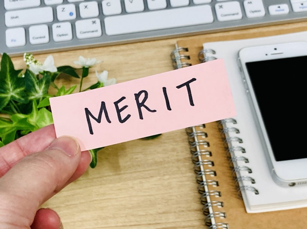 画像：「MERIT」と書かれた付箋を持つ手とキーボード、スマホ、ノート